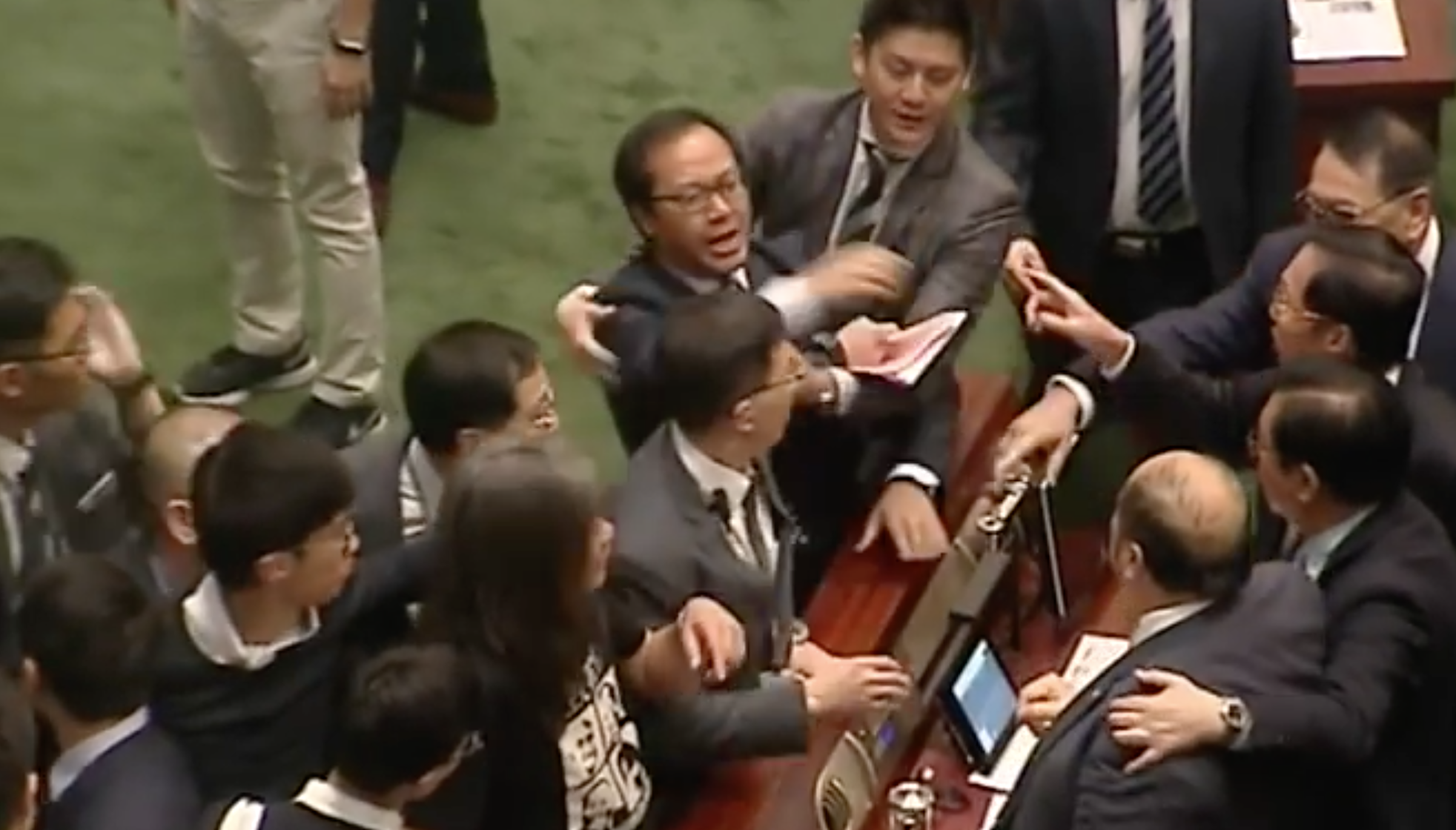2016年11月2日，香港立法會建制、非建制議員對罵期間，梁繼昌自行倒地的瞬間。用身體保護梁頌恆的梁國雄莞爾一笑。（電視截圖）